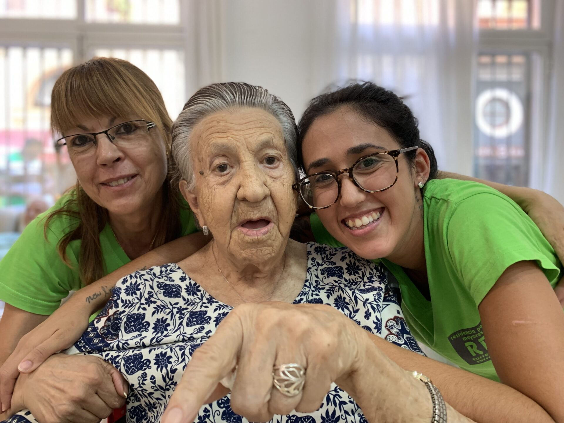 Imagen de señora mayor abrazada por dos trabajadoras de Torres Serrano apuntando a la webcam.